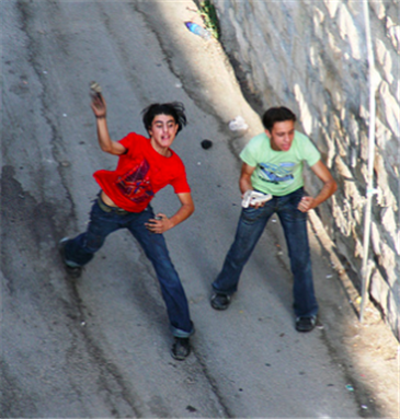 [Two Palestinian youths throw stones at an occupation military jeep. The soldiers respond by firing live ammunition. Later in the day, the boy in the red shirt was shot in the stomach and critically injured. Creative commons.]