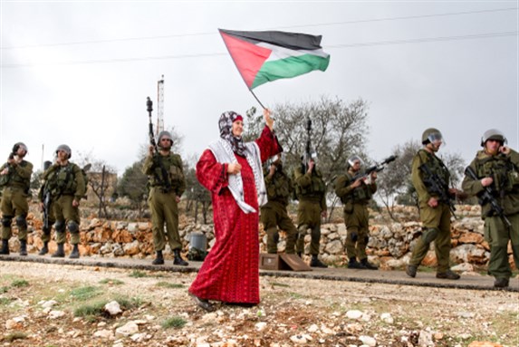 Bilin, Palestine, December 31, 2010: Palestinian woman walks with Palestinian flag in front of Israel Defence Force soldiers during weekly demonstrations against Palestinian land confiscation in Bilin. Image via Shutterstock.