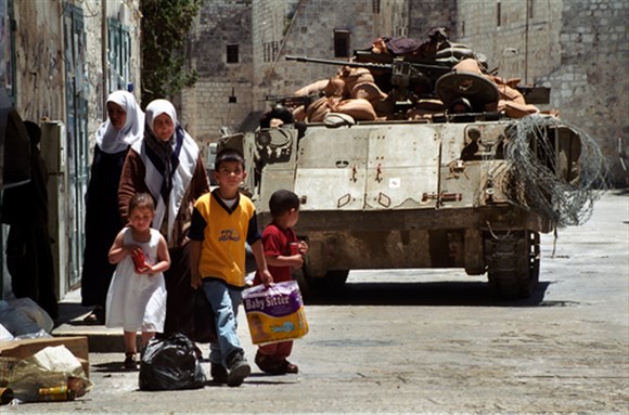 Palestinian residents of Bethlehem walk near an Israeli tank guarding Manger Square during a lull in curfew imposed by the Israeli military May 28, 2002 in Bethlehem. Image via Shutterstock.