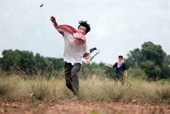 A male teenager wearing a keffiyeh to cover his face is throwing stones while his friend waving a Palestine flag at the background. Image via Shutterstock.