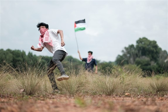 A male teenager wearing a keffiyeh to cover his face is throwing stones while his friend waves a Palestine flag at the background