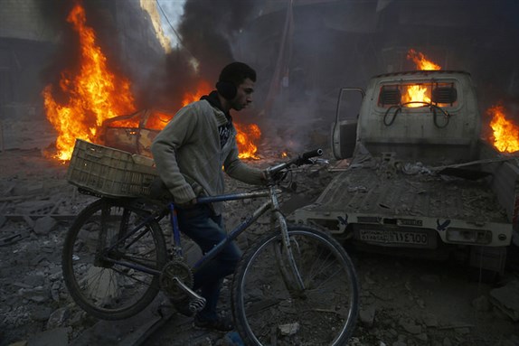 [Image of a Syrian man carrying his bicycle through the flames of Hamouria (east of Damascus). Image by Camera Jabber via Flickr]