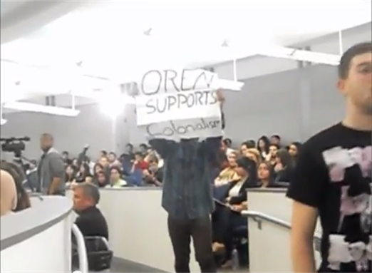 [Student participating in the walkout carries a sign reading \"Oren Supports Colonialism\". Image from video below.]
