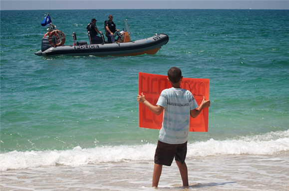 [A child carring a sign that reads `massacre on high-seas` stands on the beach-front in Ashdod. Photo by Edo Medicks used under a Creative Commons license.]