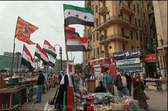 [People walking past Syrian and Egyptian flags in Tahrir Square in Cairo on 19 February 2012. Image originally posted to Flicker by Freedom House]