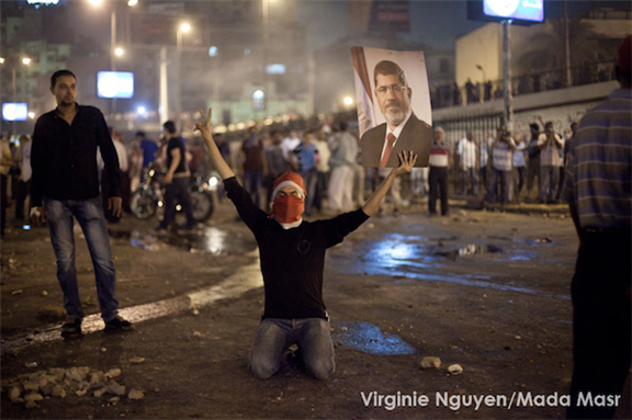 [Morsi supporter holds a poster of Mohamed Morsi in one hand and referring to \"victory\" with his other hand. Image originally posted to Mada Masr, taken by Virginie Nguyen.]
