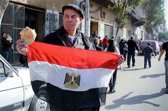 [25 January 2013, protester holding a piece of bread and Egypt’s flag in commemoration of the second anniversary of the January 25 Revolution, in which it broke out in 2011 under one unified popular demand; “Bread, Freedom, and Social Justice.” Image originally posted to Flickr by Gigi Ibrahim.]