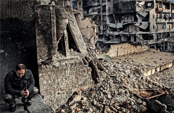 [Image of a sergeant in Lewa Salaheddin sits in front of a block of destroyed buildings in Aleppo, Syria. Image by Patrick Tombola/Laif via Redux] 