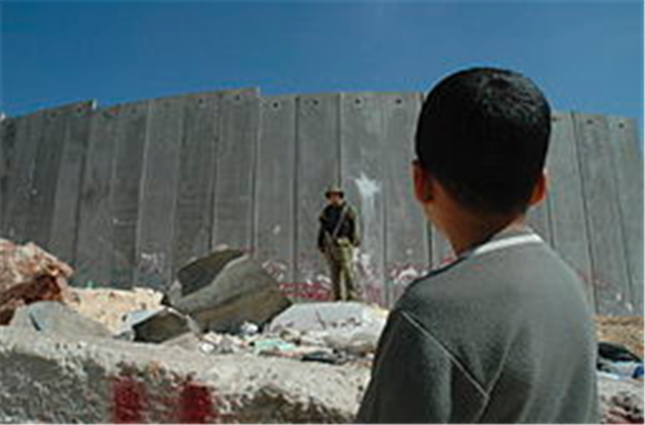 [A Palestinian boy and Israeli soldier in front of the Israeli West Bank Barrier. Picture taken by Justin McIntosh, August 2004. Uploaded from Wikimedia Commons.]