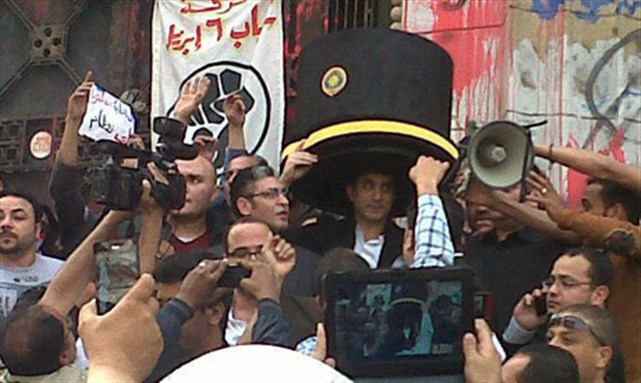 [Egyptian TV host Bassem Youssef arrives at high court on Sunday wearing a hat that pokes fun at President Morsi`s visit to Pakistan where he wore a similar hat at an honorary doctoral induction in Pakistan last month (Photo: facebook.com/11FebFront)]