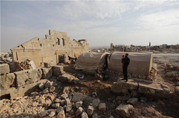 Mohamad Othman, 30, stands with his children beside their tent in the archaeological site of Sarjableh, in the northern countryside of Idlib, Syria. [Khalil Ashawi/Reuters]