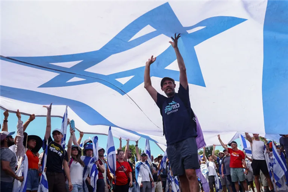 Protesters hold up a large Israeli flag at a demonstration following a parliament vote (Jerusalem, 24 July 2023). Photo via Al Jazeera (Reuters/Ronen Zvulun).