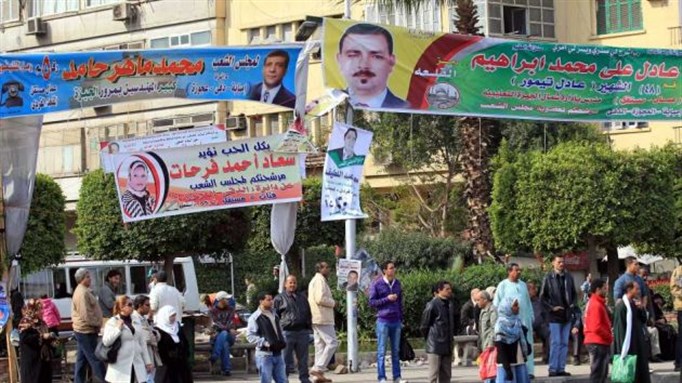 [Egyptians walk beneath electoral banners and posters ahead of the second round of parliamentary elections, in Giza, Egypt, 13 December 2011. The second round of voting will be held on 14 and 15 December in nine governorates. Image from EPA]