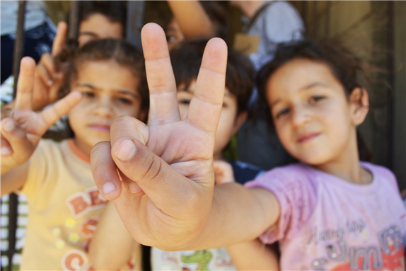 Syrian refugee children at a half-built apartment block near Reyfoun in Lebanon, close to the border with Syria, give the peace sign. The families fled Syria due to the war and are now living on a building site. (Photo: Eoghan Rice) via Wikimedia Commons